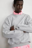 Mannequin portant un Sweat Crewneck Gris LATER... avec une chemise rose et blanche rayée en dessous, bras croisés.