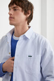 Surchemise ÉPURE Marine | LATER... rayée bleu ciel portée par un homme sur un sweat bleu. Détail vert sur la poche poitrine.
