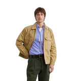 Homme portant un Blouson Worker beige, une chemise rayée bleue et rose, un pantalon plissé vert olive et des chaussures noires.