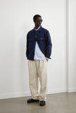 Homme portant le Blouson Worker Marine LATER..., chemise rayée bleue, pantalon plissé beige, mocassins noirs et lunettes de soleil.