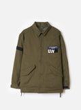 Parka CR Raf Jacket Universal Works olive, avec boutons, poches à rabat et patchs noirs sur la poitrine et la manche.