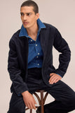 Homme en veste et pantalon en velours côtelé bleu marine Morgan 50004 NN.07, chemise denim, assis sur un tabouret.