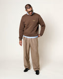 Homme portant un sweatshirt MKO Logo Cocoa HOMECORE marron, chemise bleue, pantalon plissé beige et chaussures noires.