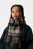 Femme portant une écharpe Carhartt WIP Parson Scarf WIP Dusty H à carreaux, couleurs marron, gris et noir, avec franges.